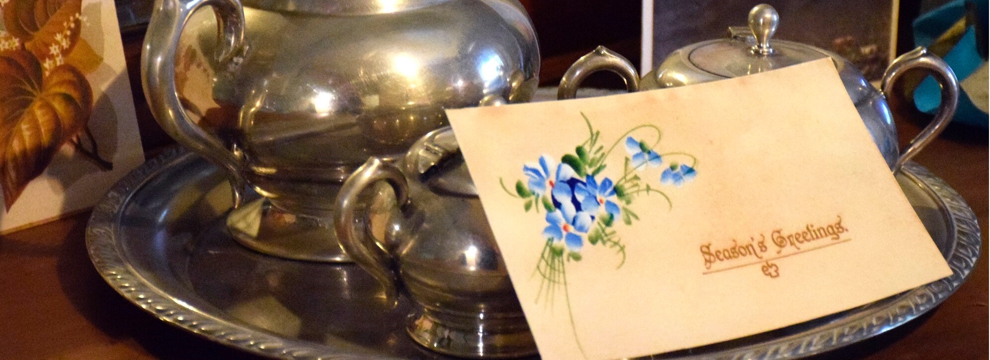 cropped image of silver teapot with old card leaning against it with the words 'Seasons Greetings' and a blue flower in the corner. Part of the Christmas at the Old Treasury Building 2018 display