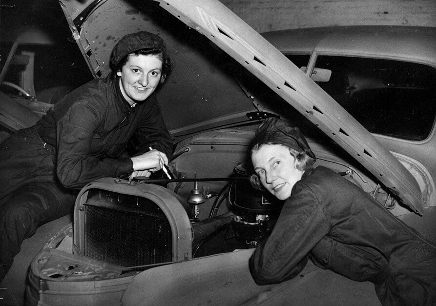 Two women (D. Austin and D. Pelling) looking under the hood of a car. Both wear boiler suits and have scarves wrapped around their hair.