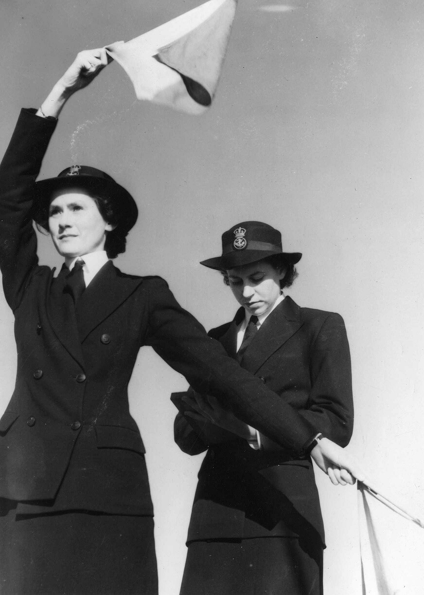 A Woman in the foreground is making signals with flags (one in each hand). Behind her a woman makes notes in a book. Both are wearing dark military uniforms with wide brim hats. They are identified as JC Barnes and DJ Eaton.