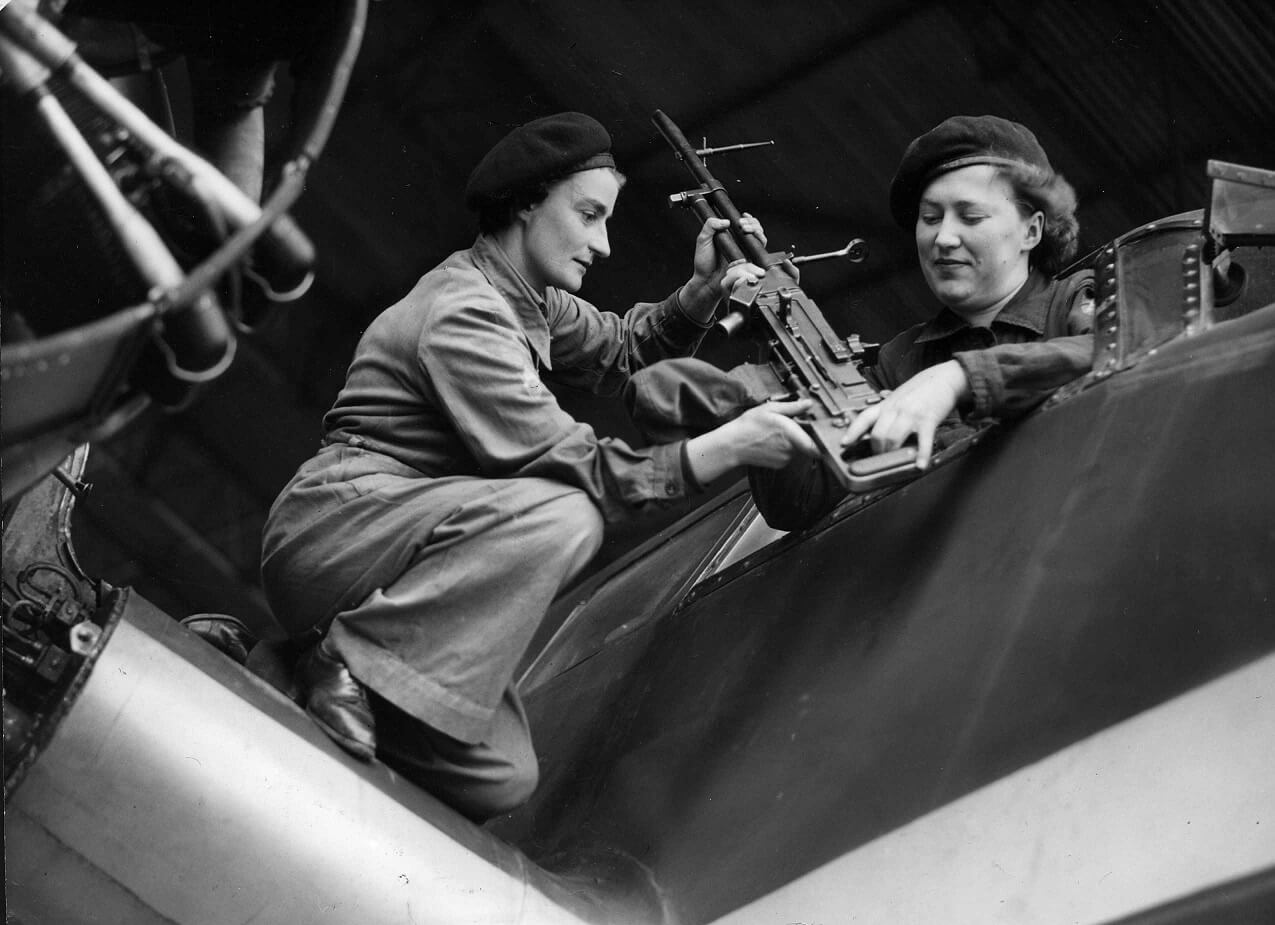 Margaret Deal and Rosemary Kemp attach a gun to the side of an aeroplane. Both are wearing boiler suits and berets.