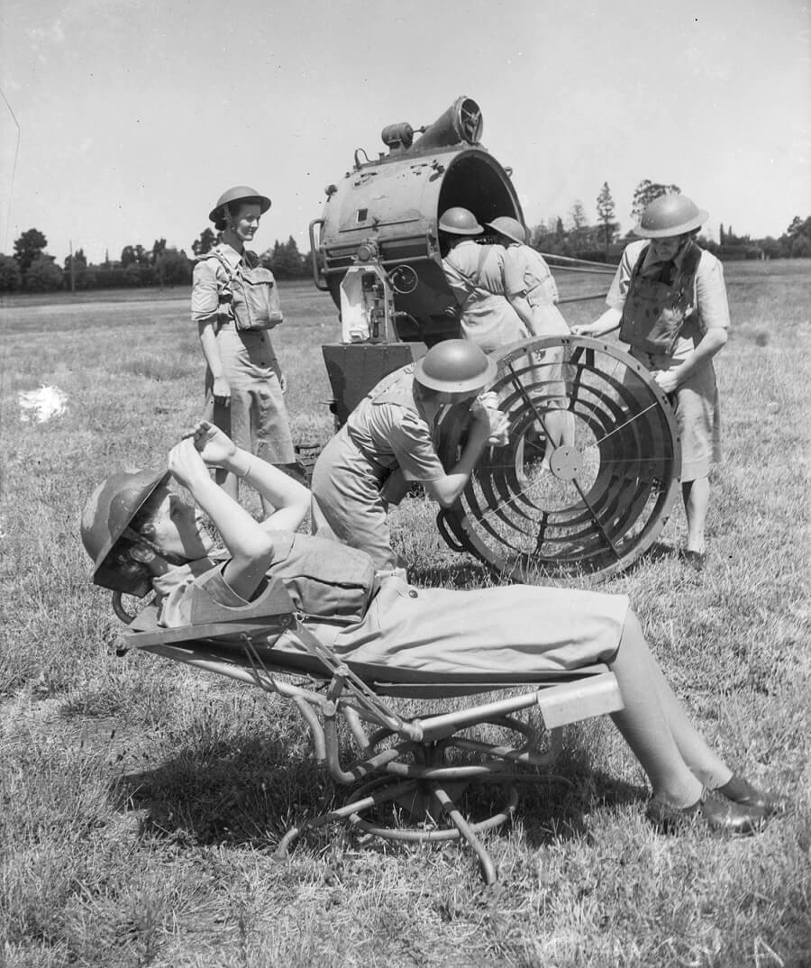 In the foreground, a woman lies on a relined chair looking up to the sky using binoculars. Behind her 5 women are inspecting or repairing a large searchlight. All are wearing a uniform consisting of a dress and metal helmet.