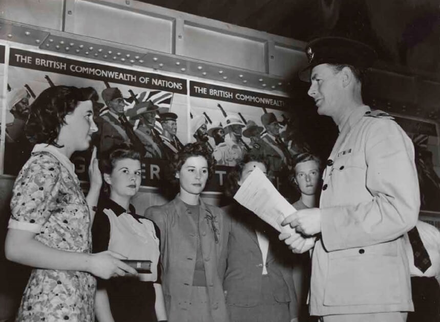 A line of women with a military appearing man standing in front of them holding papers. The first woman in line (Miss Shirley Beckett) is holding a book (possibly the bible) and has her left hand held as if giving an oath.