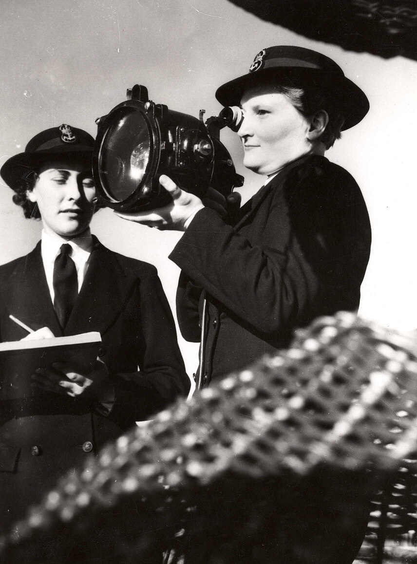A woman holds a searchlight to her eye, while another stands beside her holding a pen and clipboard. They are identified as S Flower and M Walker.