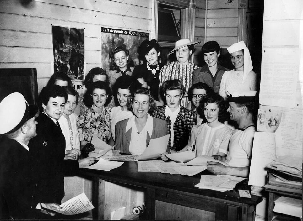 15 civilian women behind a table holding papers with a male naval officer.