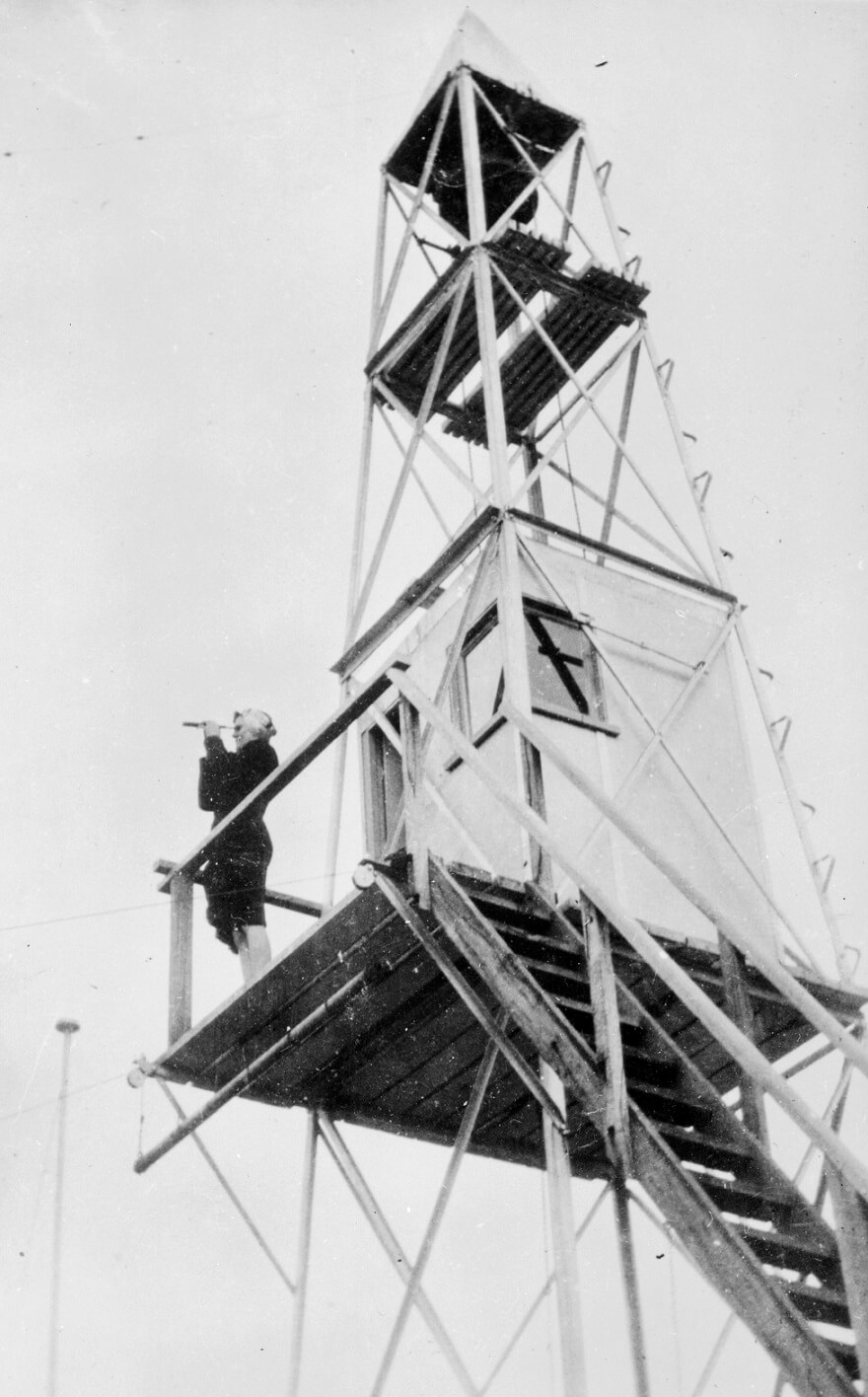 black and white photograph shows a tall simply constructed fire tower with narrow stairs leading up to a platform with an enclosed room (approx 1 metre sqaure). A woman stands in a corner of this platform near a rail with a spyglass to her eye. She is wearing a dark suit jacket and skirt. 
