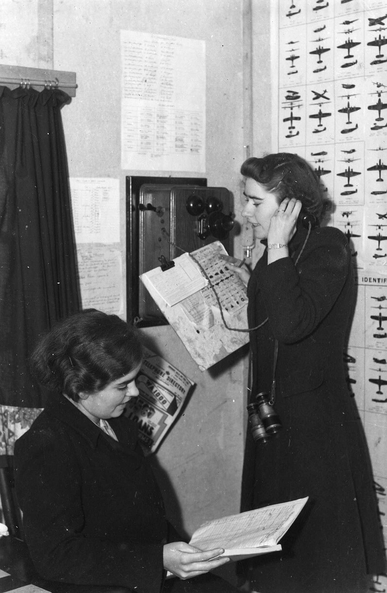 black and white photograph showing two women wearing identical dark dresses. One to the right of the image is talking on a telephone while holding a chart. The second sits to her left reading from a paper notebook. On the wall behind them is a chart showing various types of aircraft, with the sides, back, and top of the aircraft in silhouette. 