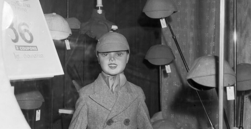 Black and white photograph showing a child (boy) mannequin wearing a double breasted coat in a shop window display. A sign near the mannequin announces it as for sale for 3 coupons.  