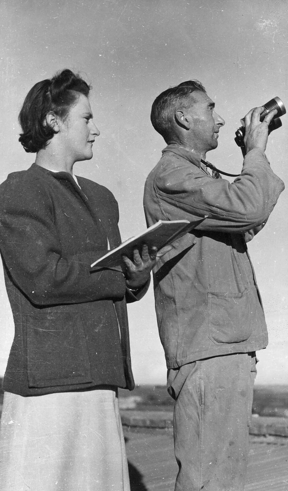 Black and white photograph shows 2 people. On the left is a woman with short curled hair. She is wearing a dark jacket over a pale skirt, and holding a paper notebook open. To the right is a man holding binoculars as if putting them to his eyes, or taking them away. He is wearing worn work-mans jacket and pants. 