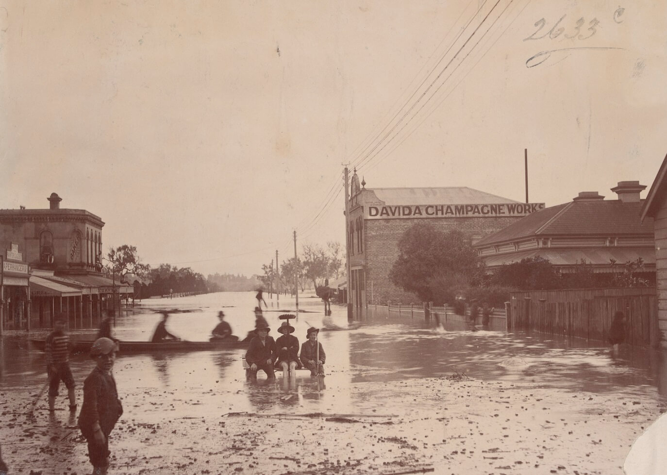flooded street with houses on either side. A large building on the right side of the image advertises "Davida Champagne Works". 3 people  sit on a bench in the flood water, while another 4 row past. A young boy stares at the camera from the left side of the image.