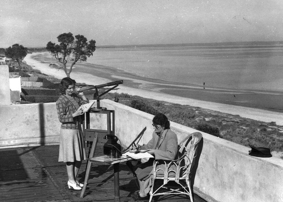 black and white photograph showing a balcony by the seaside. Two women are working, the one on the left is holding a small telescope while examining what looks like a map. She is wearing a pale swing skirt and checked cardigan. Her companion is seated in a cane chair writing in a book. One of their hats sits on the ledge of the balcony.  