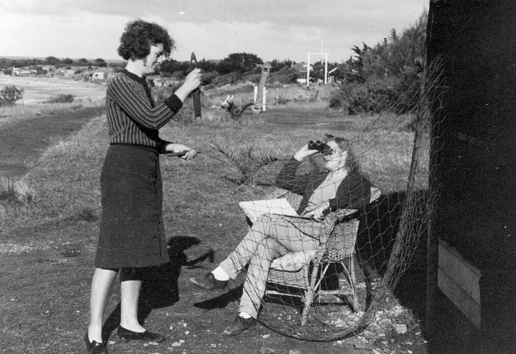 Black and white photograph shows 2 women. They are in a rural setting, surrounded by glass, there may be a shore line with beach behind them. One is standing, wearing a striped jumper with a dark pencil skirt. The is holding an implement and repairing a net with large (approx. 10cm) holes. The second woman is sitting in a wicker chair wearing a shirt, dark cardigan and light coloured fitted trousers. She has her right hand holding binoculars to her eyes while she looks up to the sky. 