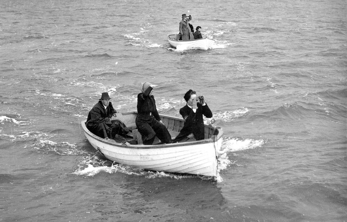 black and white photograph shows 2 boats on slightly choppy water, one is closer to the view, the other further away. The closer boat has 3 passengers, a man at the rear wearing an overcoat and hat, a woman slightly in front of him dressing in coveralls and a knitted scarf wrapped around her head, she is shielding her eyes as if from the sun. The final woman sits at the front of the boat with an overcoat and gloves, staring ahead using binoculars.  The far boat appears to have 3 people also, a man in a trench coat and hat is standing up obscuring the 3rd occupant. He has his hand shielding his eyes staring forward. The second occupant is wearing a dark coat and sitting.  
