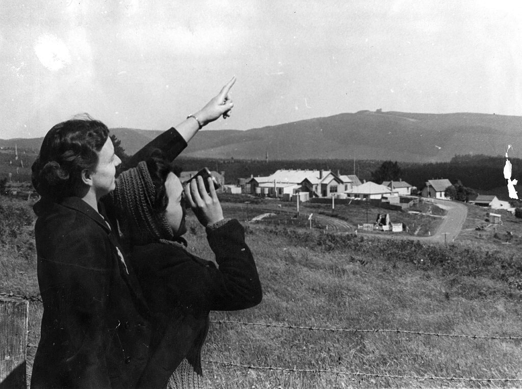 black and white photograph shows two women to the left of frame examining the sky, one woman points upwards, while the other uses binoculars. They are in a rural setting, surrounded by grass and hills and a small village in the background. 