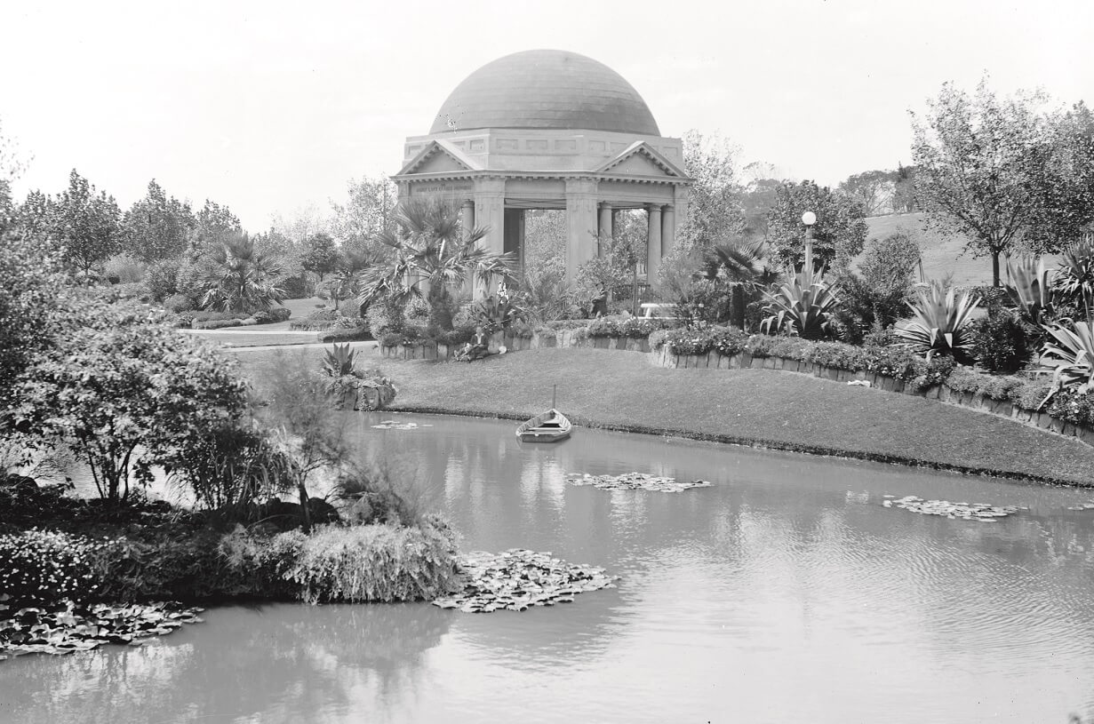 black and white photograph showing a garden with curved pond. In the middle of the image on what appears to be an island stands an elaborate though small structure. A dome over an octagonal shaped structure with just classical styled columns holding up the roof. 