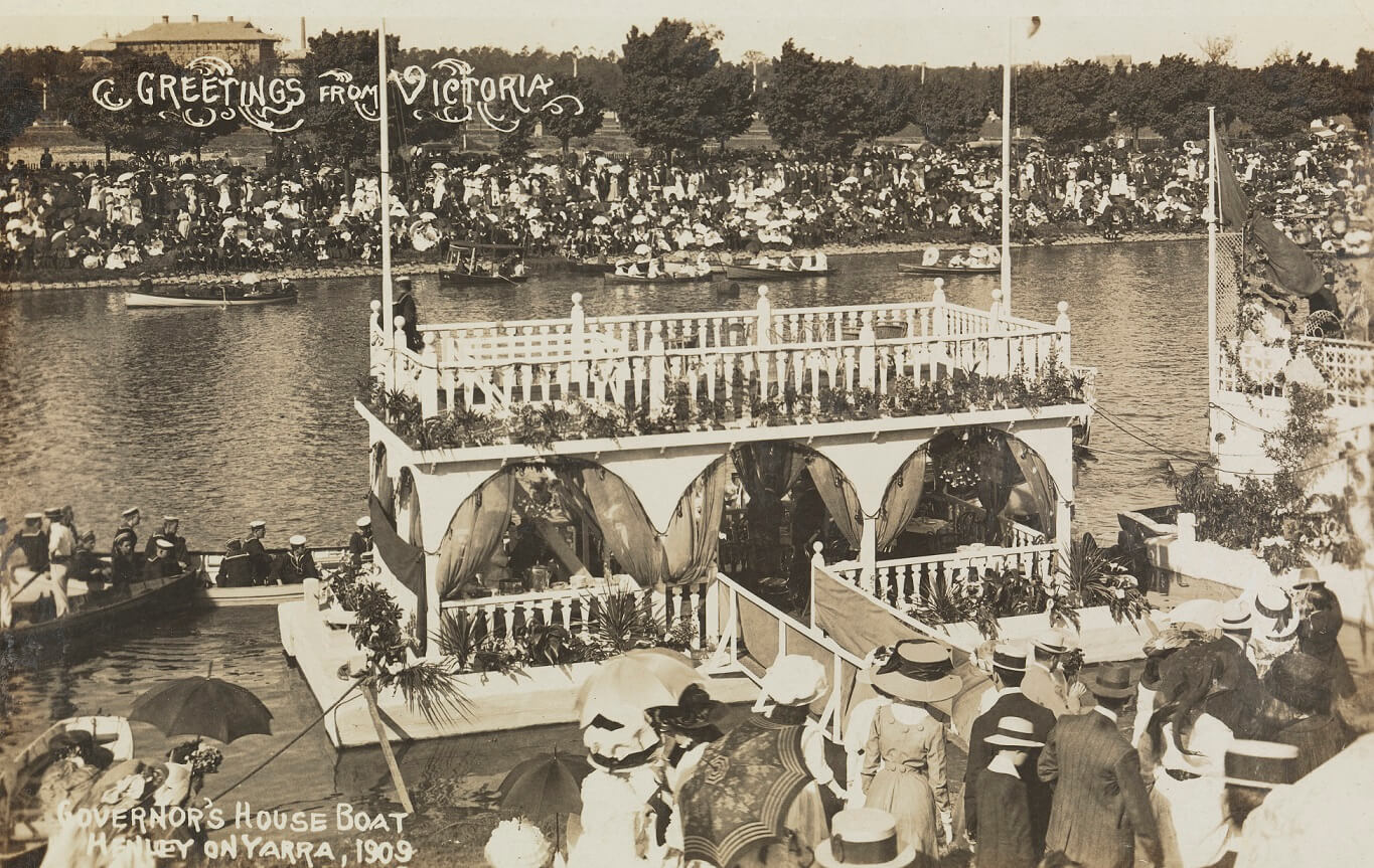 black and white photograph showing an event along a river. The main focus of the image is a rectangular construction of a house boat. It is 2 stories, with the lower level that appears to be one room, the sides of which are simple archways with billowing curtains attached to the columns. the upper level is a simple balcony with a picket fence surrounding the edge. A person stands on the upper deck. Written in the top left corner of the image is "Greetings from Victoria", and along the bottom left corner reads "Governor's House Boat | Henley on Yarra, 1909".