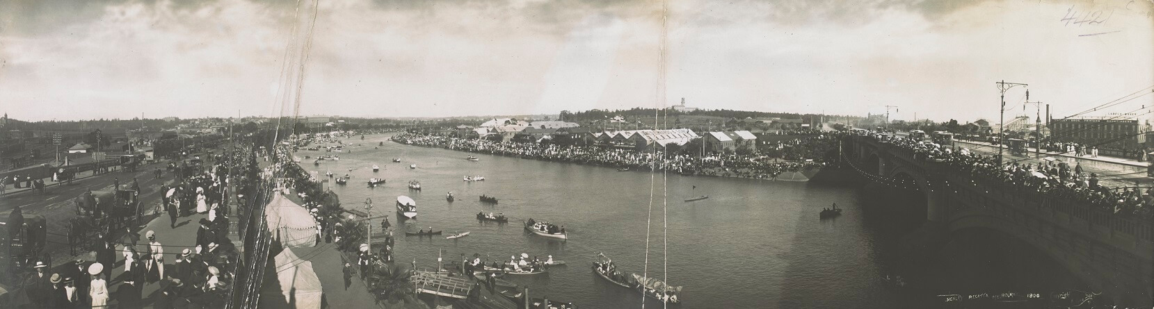 black and white image with two vertical lines indicating damage. It shows a lively event on a river. To the right of the image is a bridge crossing the river, with people lined up along the edge shoulder to shoulder. Along the left side of the river is a collection of boats, small and large (8 persons). The right bank has large temporary structures. People line both banks, all wearing hats and some carrying parasols. 