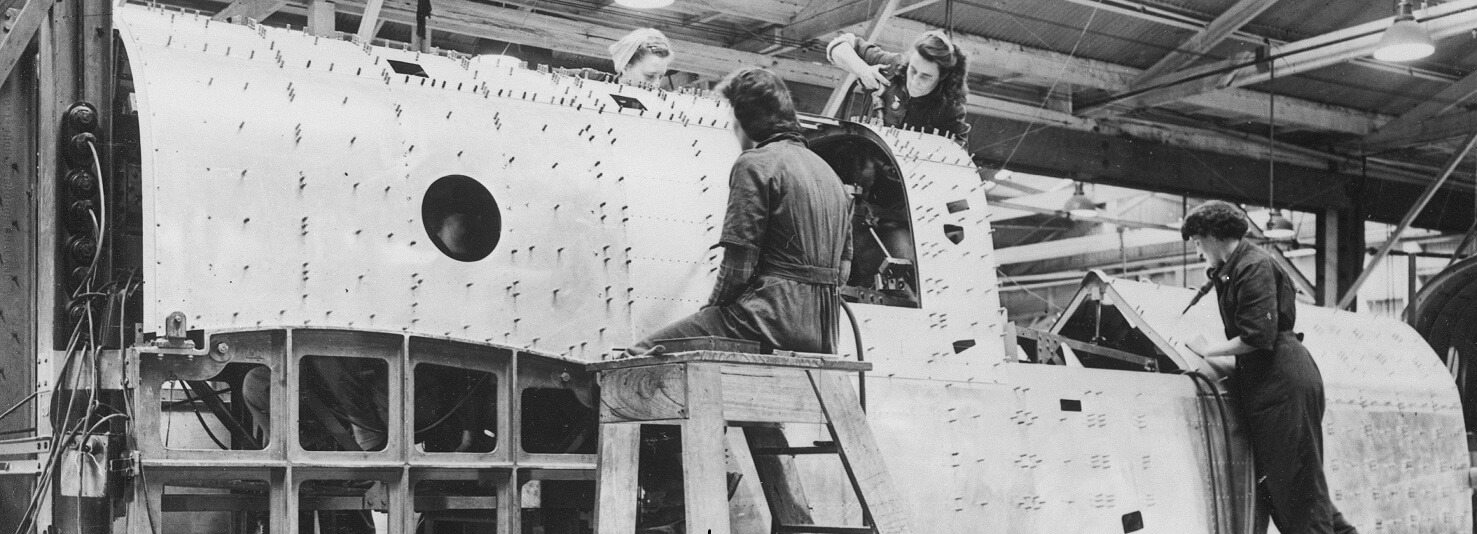 black and white photograph, 4 women are working on a large piece of metal machinery, it might be an airplane. It has rows and rows of little rivets or screws that the women are attaching. The women all wear boilersuits and have their hair in fashionable 1940s style with headscarves. 