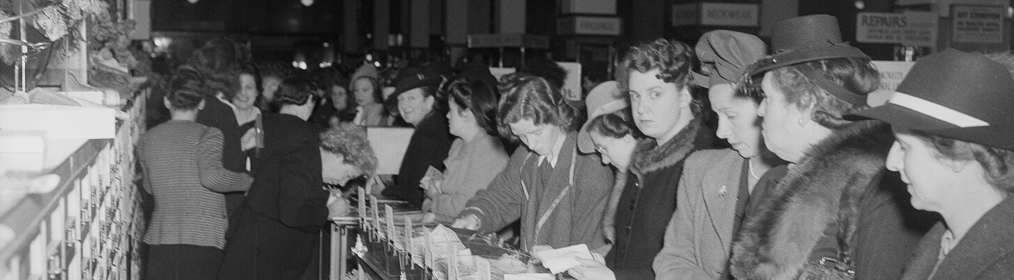 Black and white photograph of women. On the left shop assistants fuss behind a counter, on the right women in overcoats and hats line up against the counter. One woman looks straight into the camera. 
