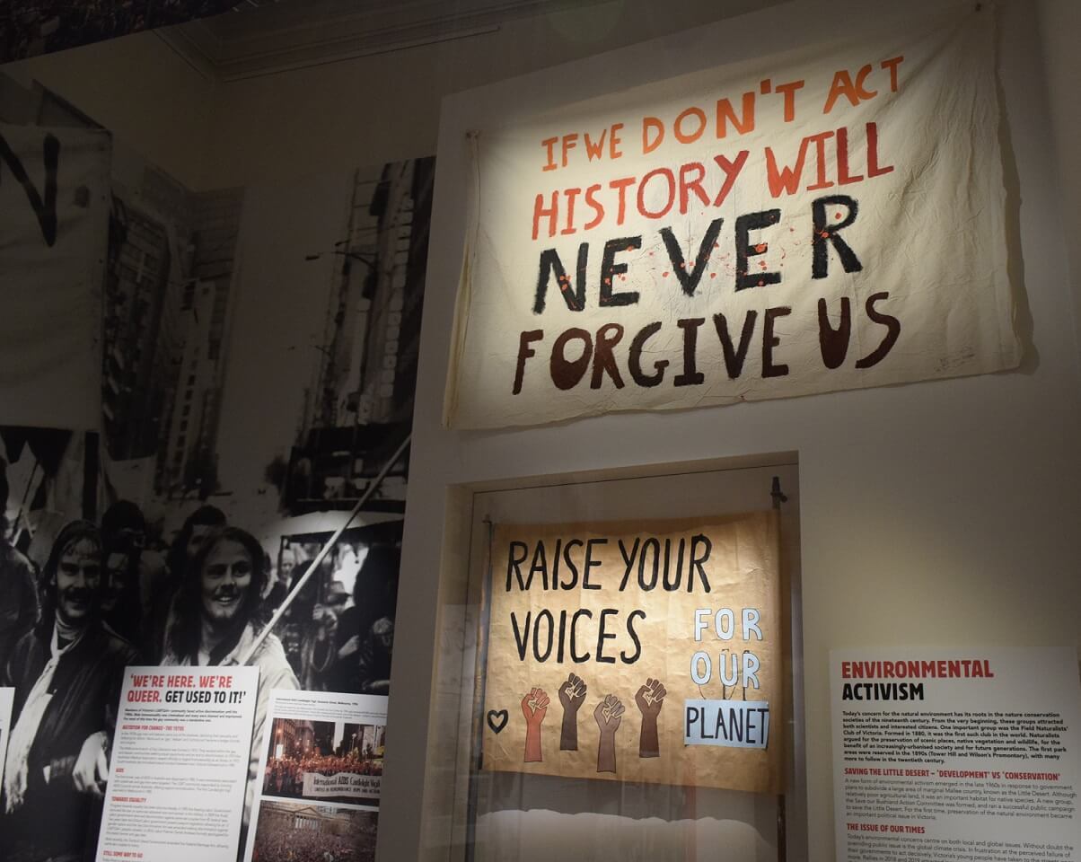 Photograph from an exhibition. The focus of the image is 2 banners displayed flat on a wall. The higher one reads "If we don't act history will never forgive us" and has red paint splattered over it to appear like blood. The lower image reads "Raise your voices for our planet" and has 4 different skin toned fists.