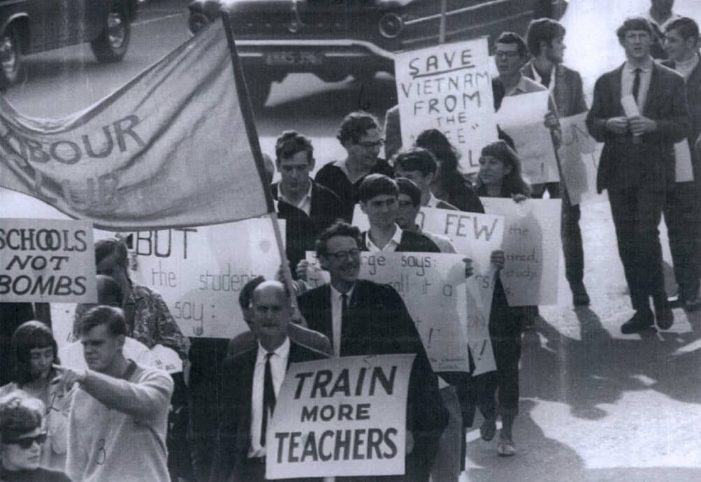 black and white image of a protest march. visible placards read "Schools not Bombs" "train more teachers" "Save Vietnam from the (unreadable)." Theres more men than women and the men are all wearing suits. A car travels beside them, like they are on the side of a road.