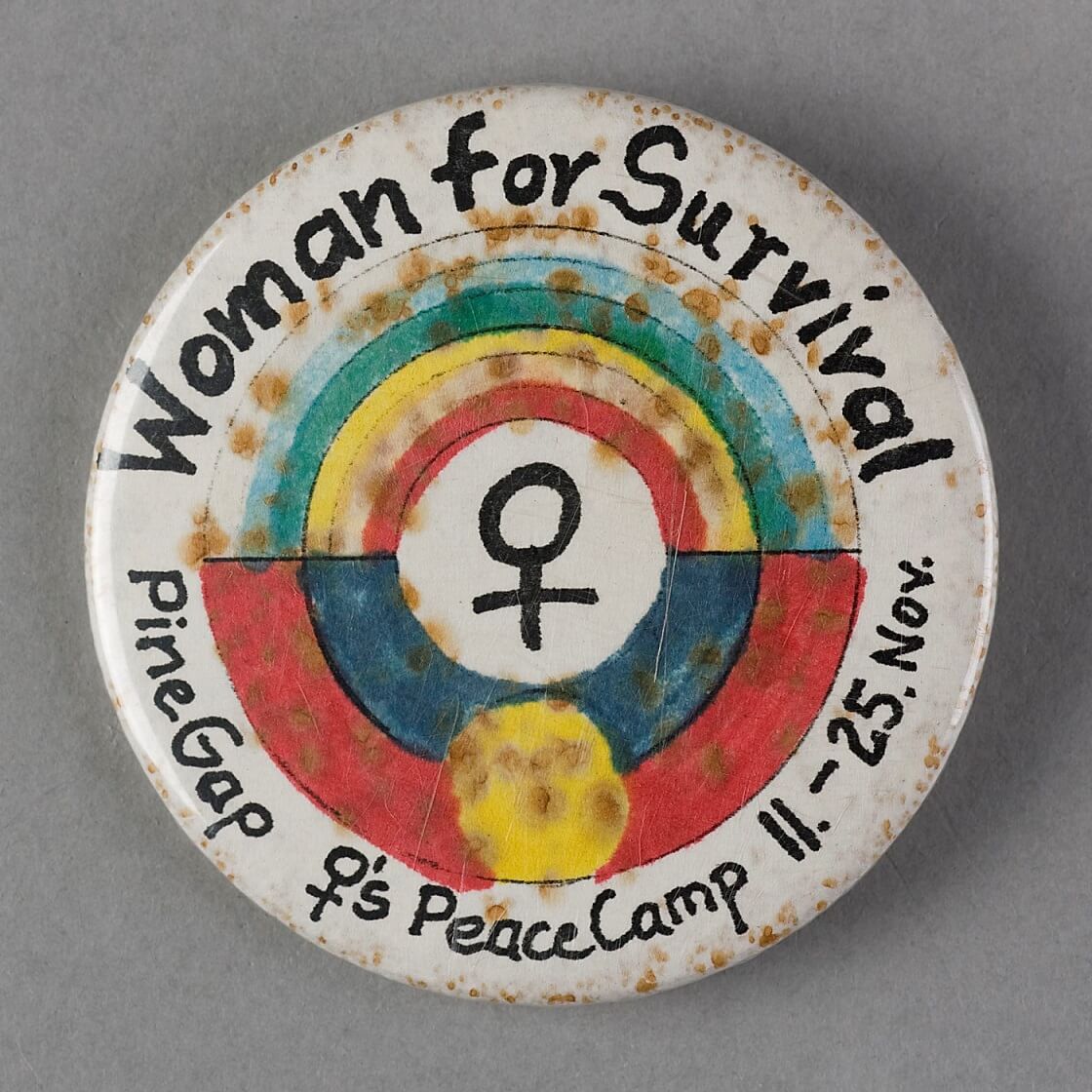 Image of a badge with text "Woman for Survival | Pine Gap Women's Peace Camp 11-25 November" with a rainbow flag and Aboriginal flag circling the womens symbol