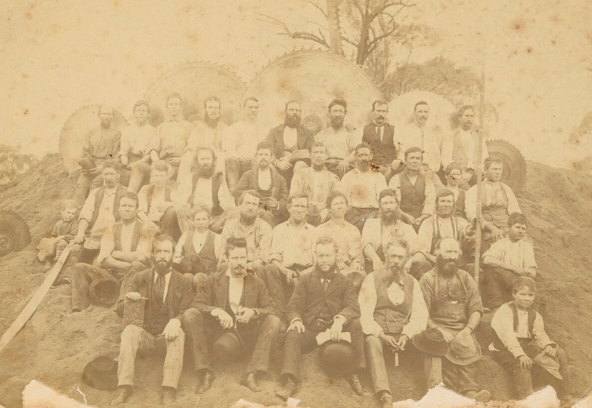 sepia toned photograph of men and young boys sitting on an embankment in 4 layers. Large circular saws are placed around them indicating their trade.