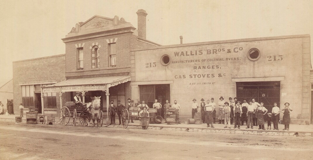 sepia toned photograph of a large business. The sign on the wall says 'Walllis Bros & Co | manufacturers of colonial ovens | Ranges | gas stoves &c" a group of men and boys stand out the front posing for the image.