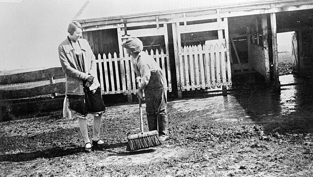 black and white photograph of a young person sweeping an outside stable.