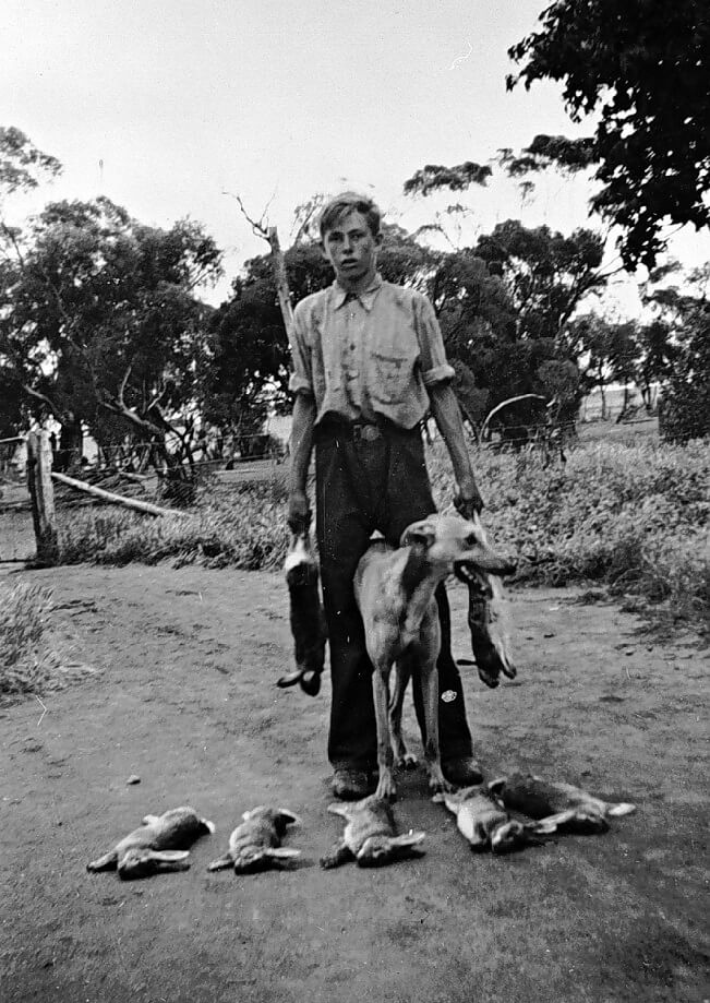 black and white photograph of a young man with a dog. he holding dead rabbits in both his hands while 5 lie at his feet.