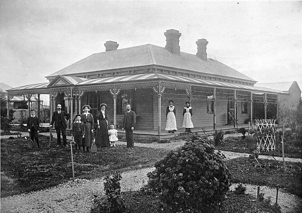 black and white photograph of a single story home with 9 people posing in a formal manner. Two of them are wearing white aprons, while the others are dressed in a more relaxed manner.