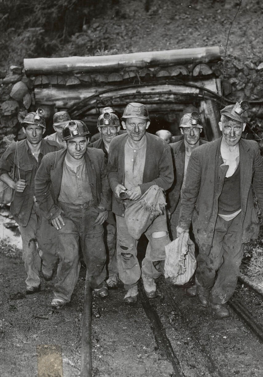 Black and white photo of men covered in coal dust. They are smiling at the camera while emerging from a mine.