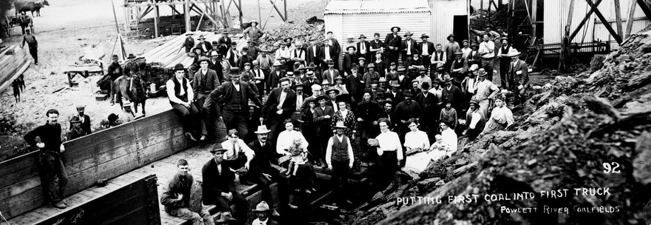 black and white photo showing approx 50 men and women smiling at the camera. hand writing over the image reads "92 | putting first coal into first truck | Powlett River Coalfields".