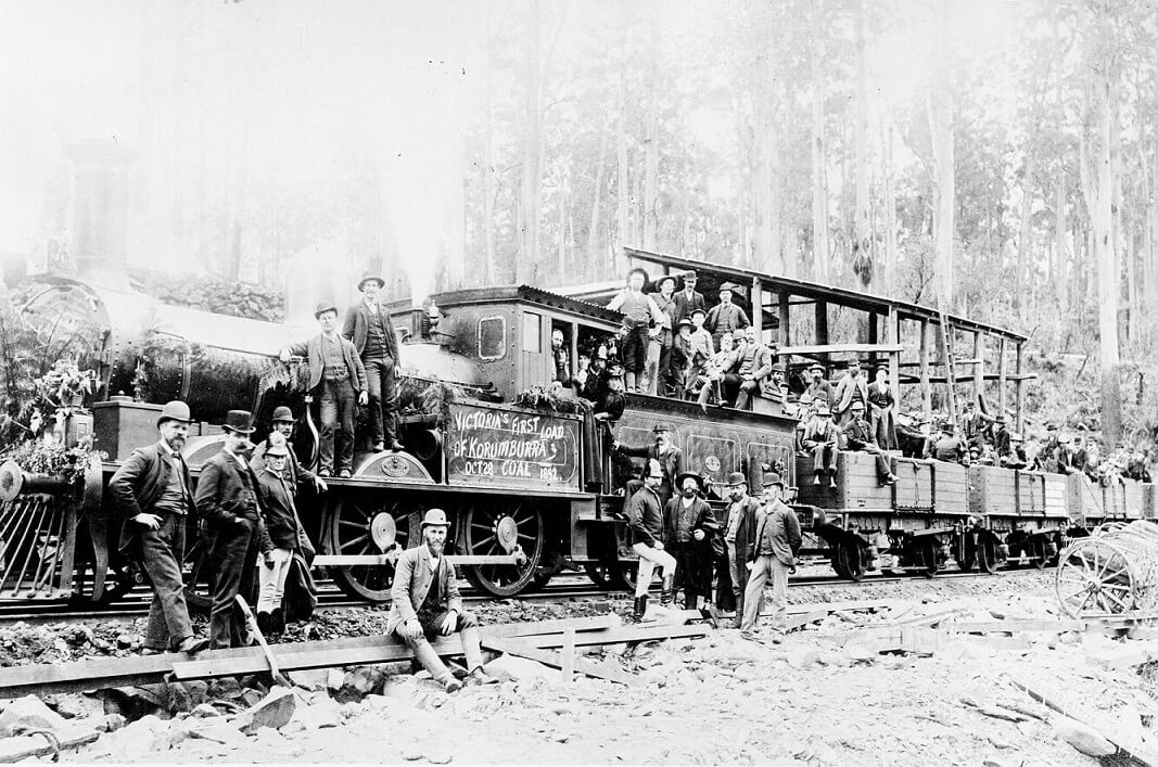 a train in a forest with men sitting on all possible surfaces. a hand-drawn sign on the side of the engine reads "Victoria's first load of Korumburra coal, Oct 28, 1892"