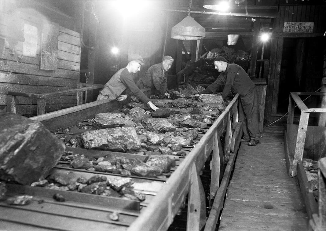 black and white photo showing a conveyor belt with coal and 3 men sorting it.