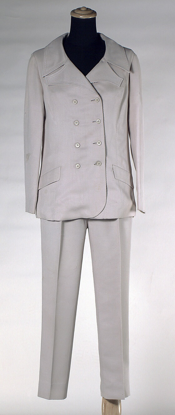 The suit consists of a twill jacket and pants. (1) Jacket is long-line tailored double-breasted with vent at the back. The short belt is buttoned to the back. The collar has sculptured revers. The front jacket features two false pockets and two rows of beige plastic buttons at fastening. The jacket is lined in beige polyester. (2) Straight legged hipster style pants with zippered front opening. Unlined.